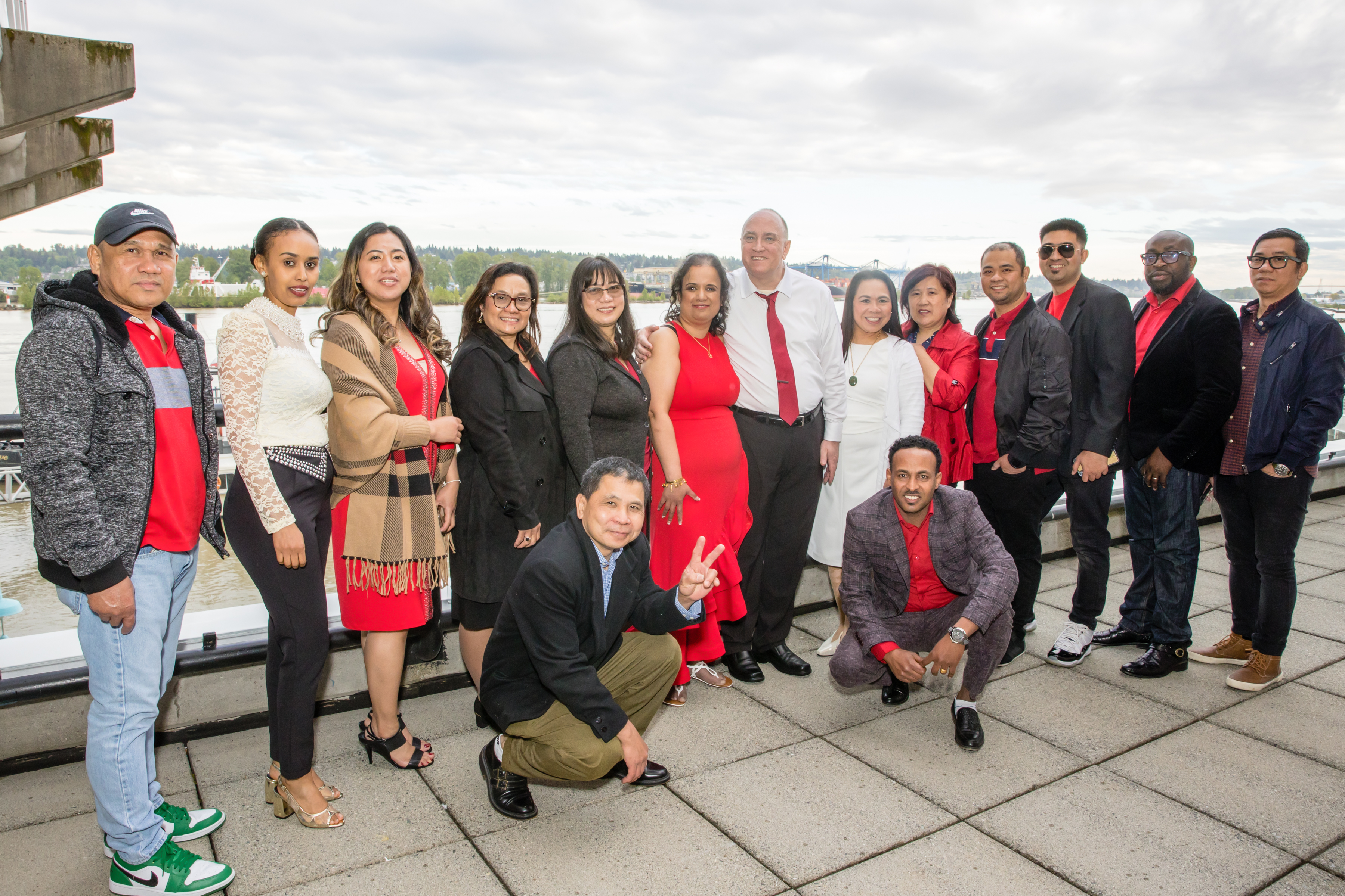 Kristy, in a long red dress, stands with a group of her students on a hotel balcony with the Fraser River behind them. Everyone is in formal wear and smiles at the camera.