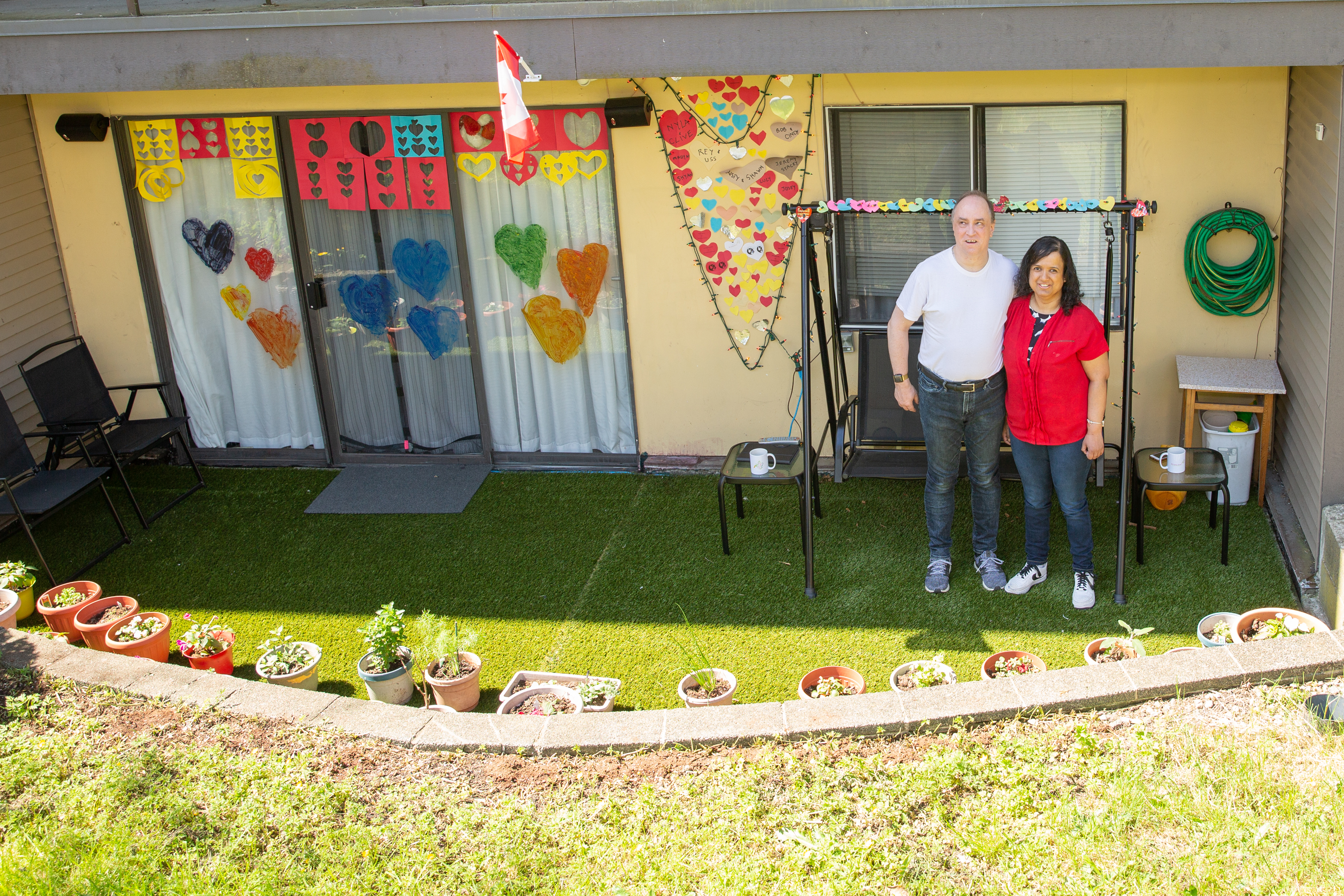 Kristy and Shawn sit on a black two-seater swing on their ground floor patio. The ground is covered with artificial grass and has dozens of flourishing flowers and herbs. On the three-panel sliding door are large painted hearts in different colours.
