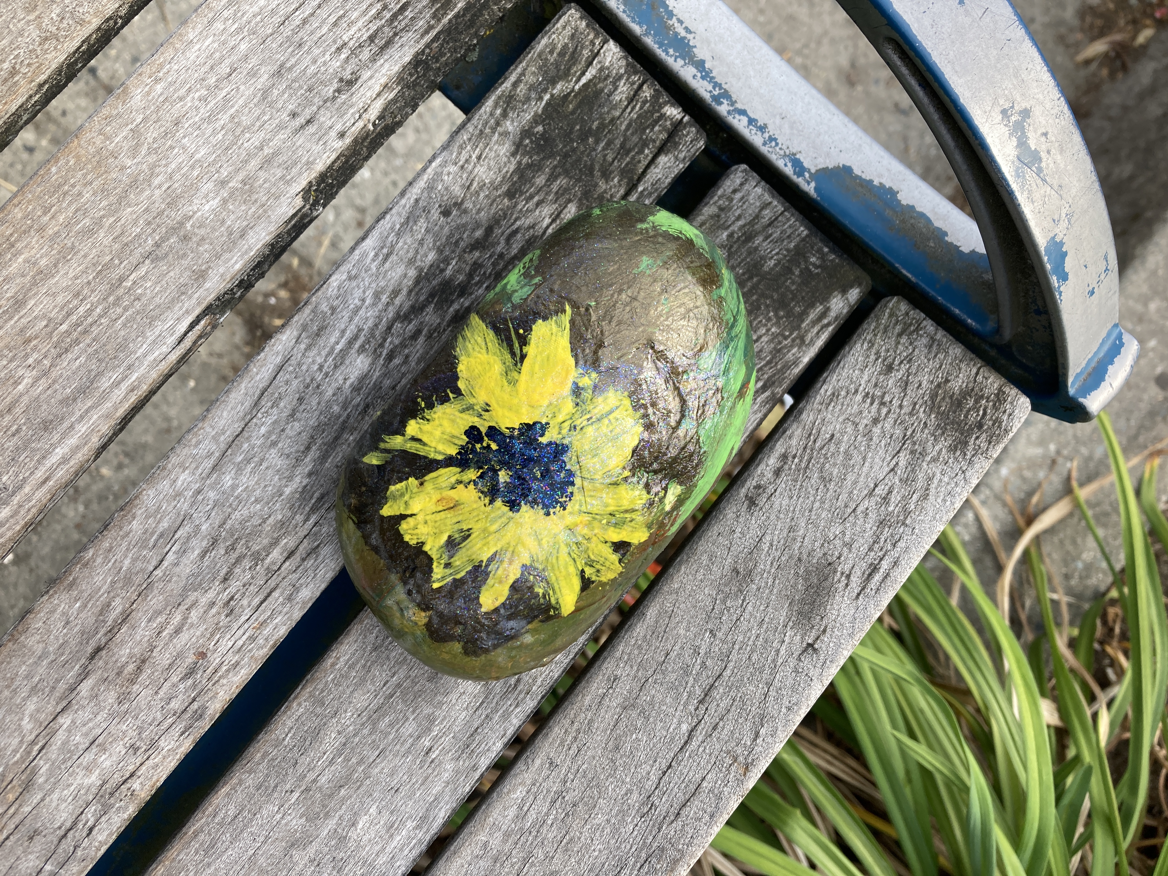 A handpainted rock, featuring a bright yellow sunflower with a dotted black and brown centre, sits on a bus bench in the sunshine.