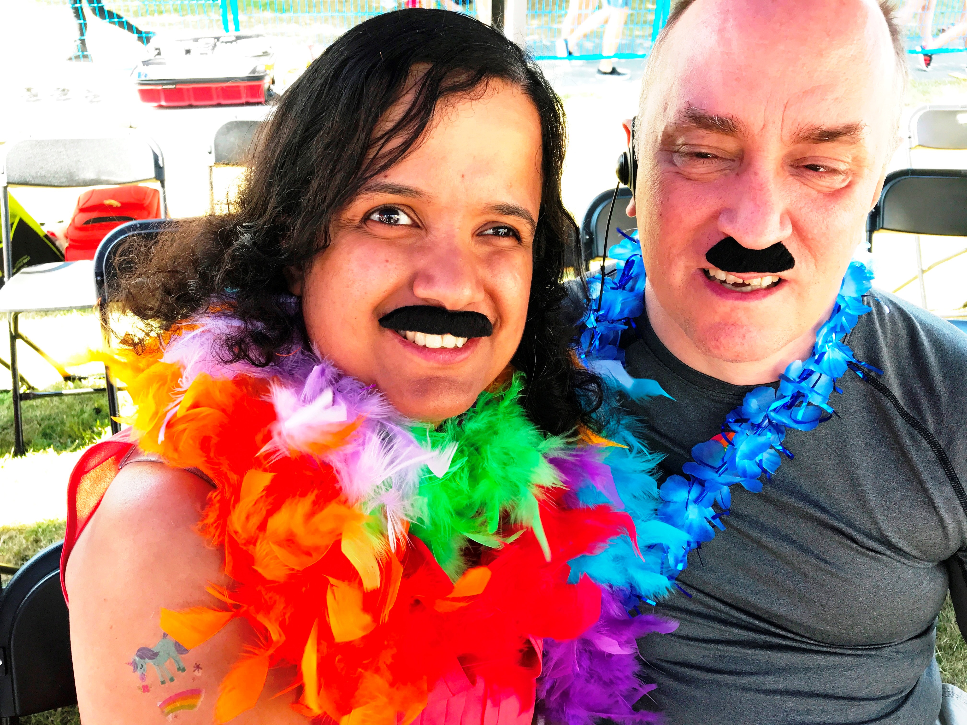 Kristy and Shawn, wearing fake mustaches and rainbow boas, under the Accessibility Tent at the Pride Parade