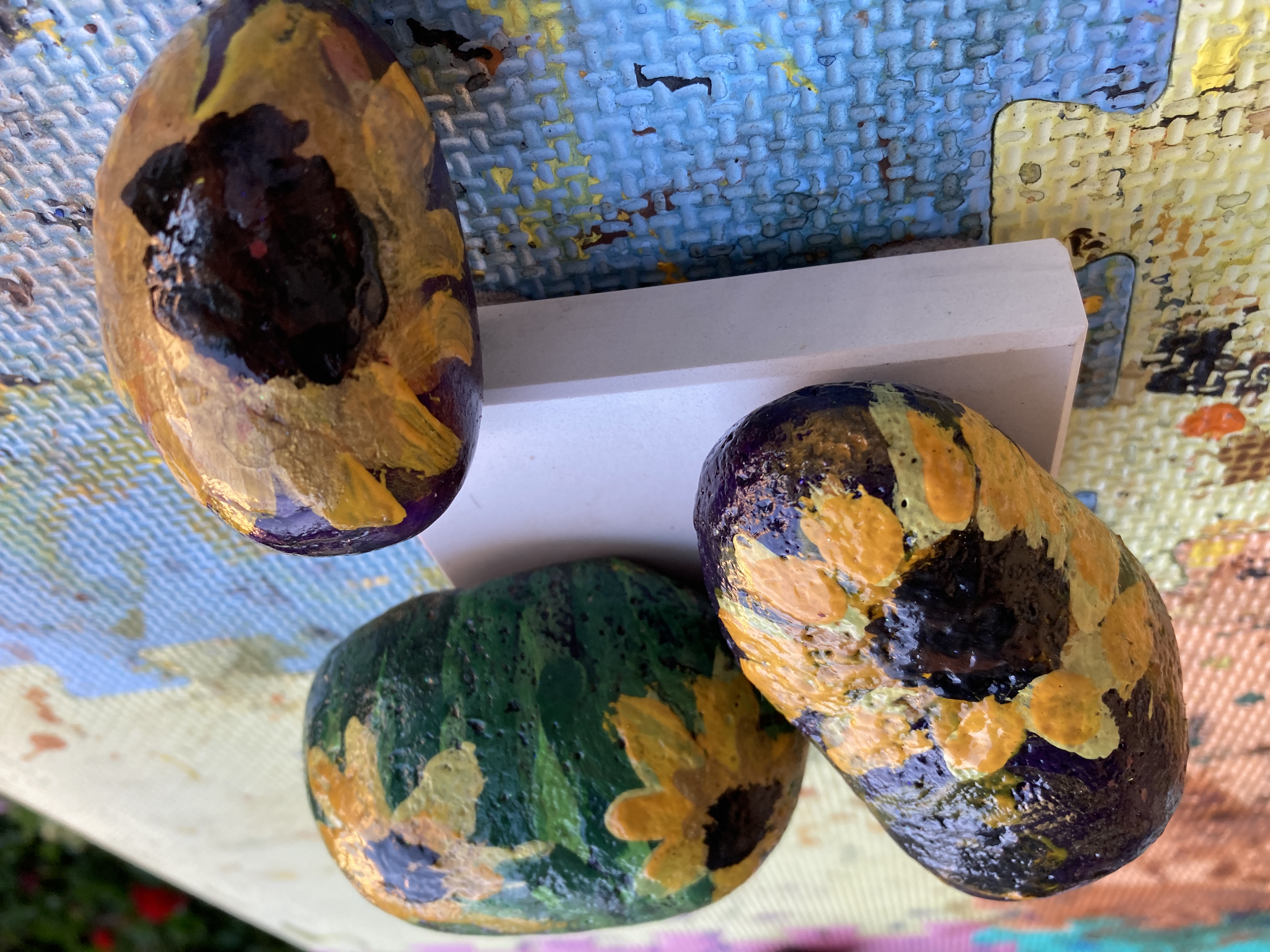 Several rocks in various sizes have solid green, blue or purple backgrounds and bright yellow sunflowers painted on them.